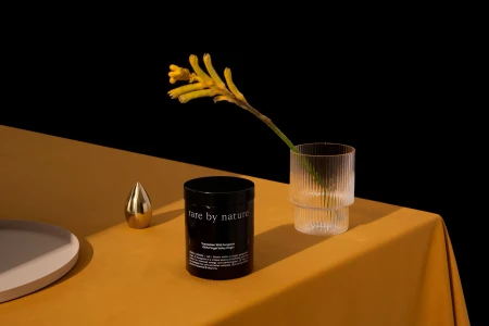 Black product jar and ribbed glass with yellow flower on a mustard cloth table against a dark background.