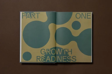 An open book displaying abstract organic green shapes and typography on mustard yellow pages against a dark brown surface
