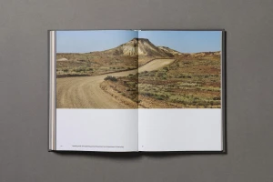 An open book resting on a neutral gray surface, displaying a landscape photograph of a winding dirt road and a barren hill.