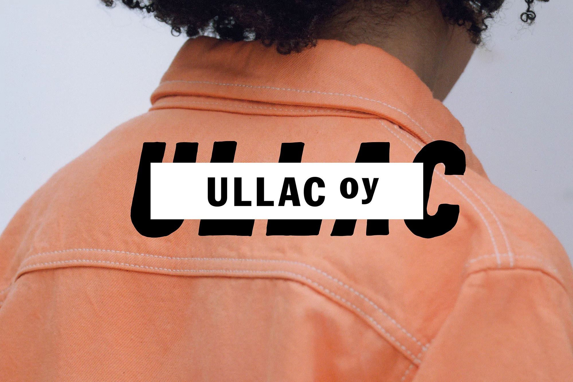 Close-up shot of an adult wearing a light-orange jacket with black ULLAC oy text printed on the back against a light-gray backdrop.