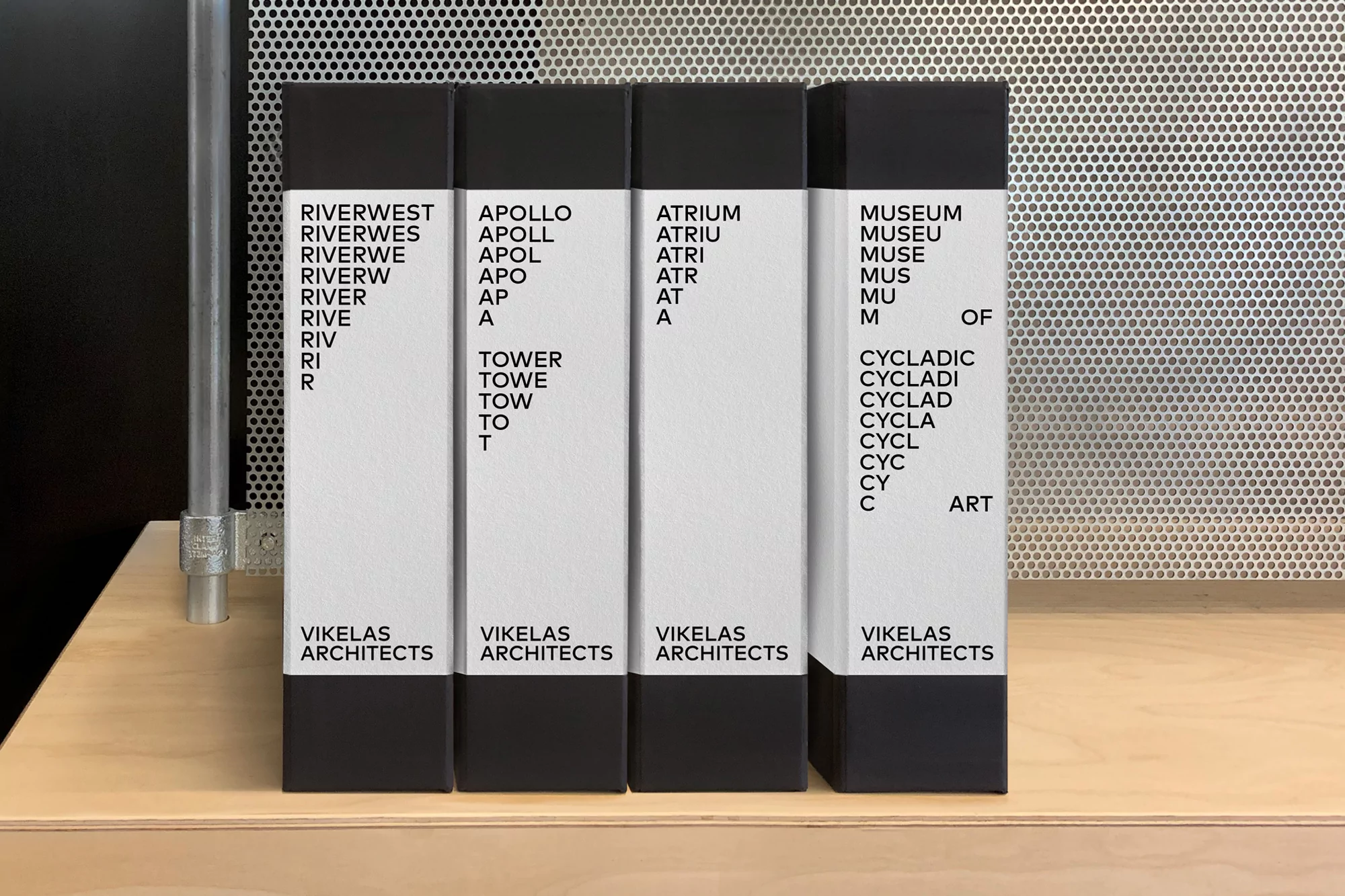 Four black and white architectural books standing upright on a light wooden shelf, showcasing Vikelas Architects branding.