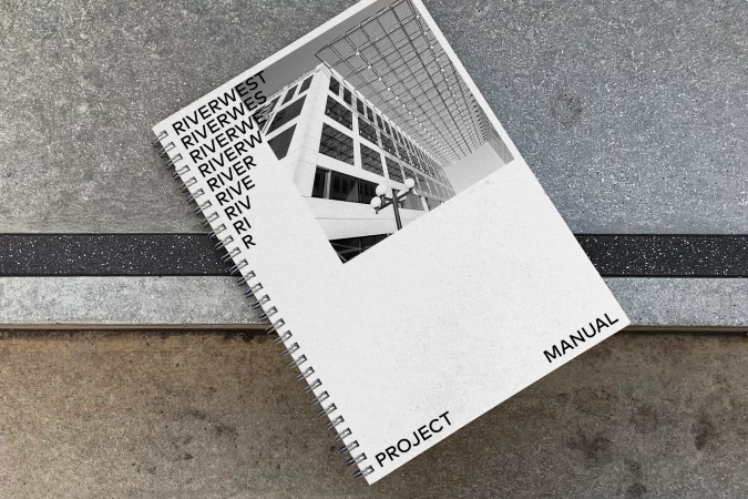 Elevated view of spiral-bound project manual featuring an architectural image, set on a textured concrete surface with a dark inlay.