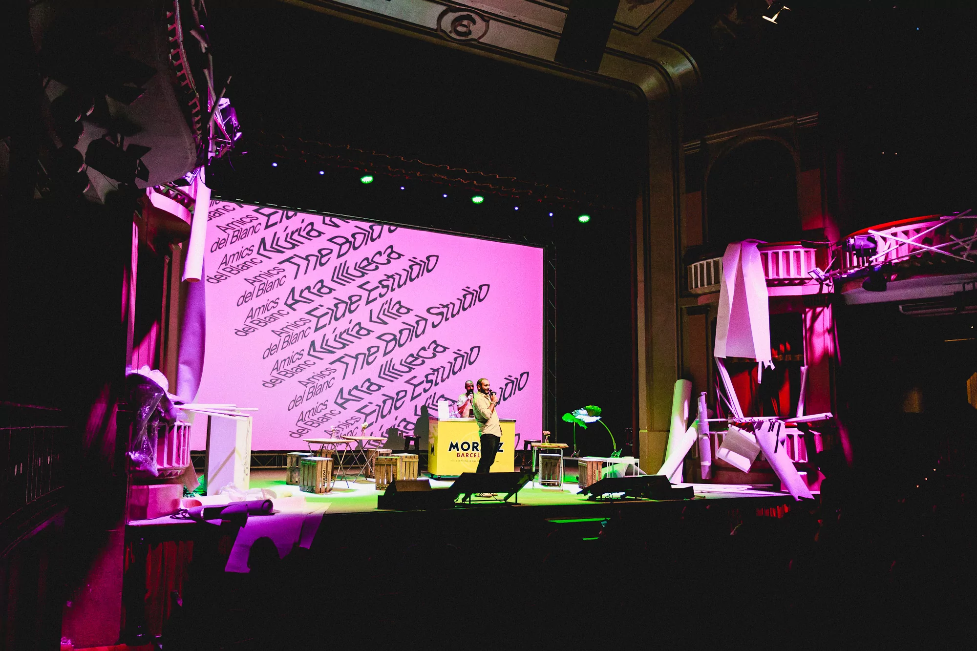 Stage view featuring a person speaking, illuminated by stage lights, with a pink backdrop and various props, creating a vibrant atmosphere.