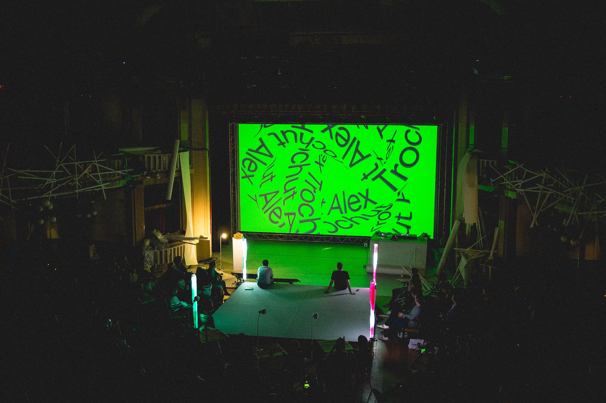 Wide shot of a stage with two people on a green screen and audience members in dark setting, with neon light props and stage decorations.