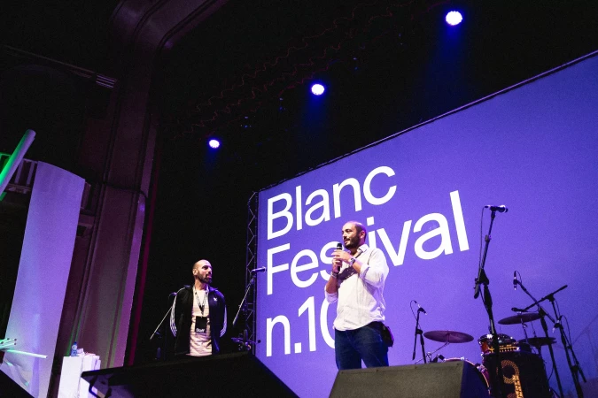 Two adult men on stage presenting at the Blanc Festival in front of a large screen with visible stage equipment in the background.