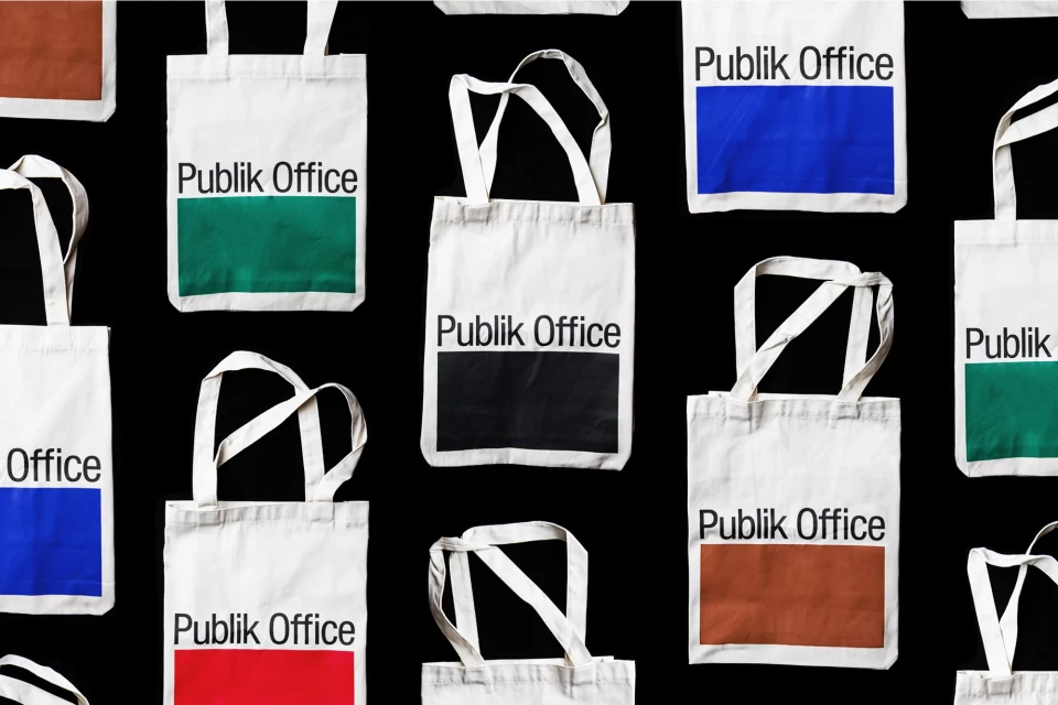 Overhead shot of multiple white tote bags with the words 'Publik Office' in black letters and a solid color block on a black backdrop.