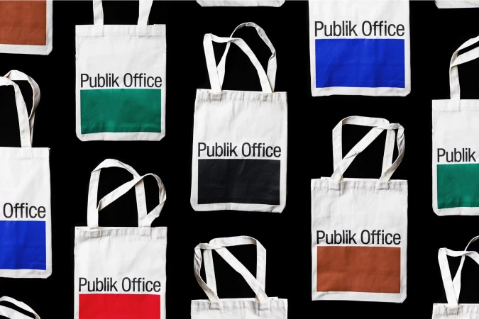 Overhead shot of multiple white tote bags with the words 'Publik Office' in black letters and a solid color block on a black backdrop.