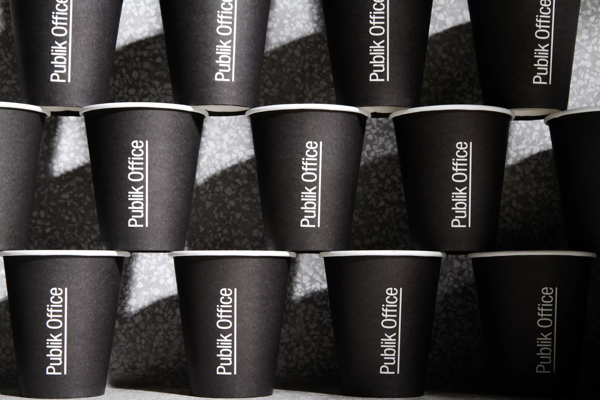 Close-up of multiple black paper cups arranged in rows and columns, each printed with the text "Publik Office" in white.