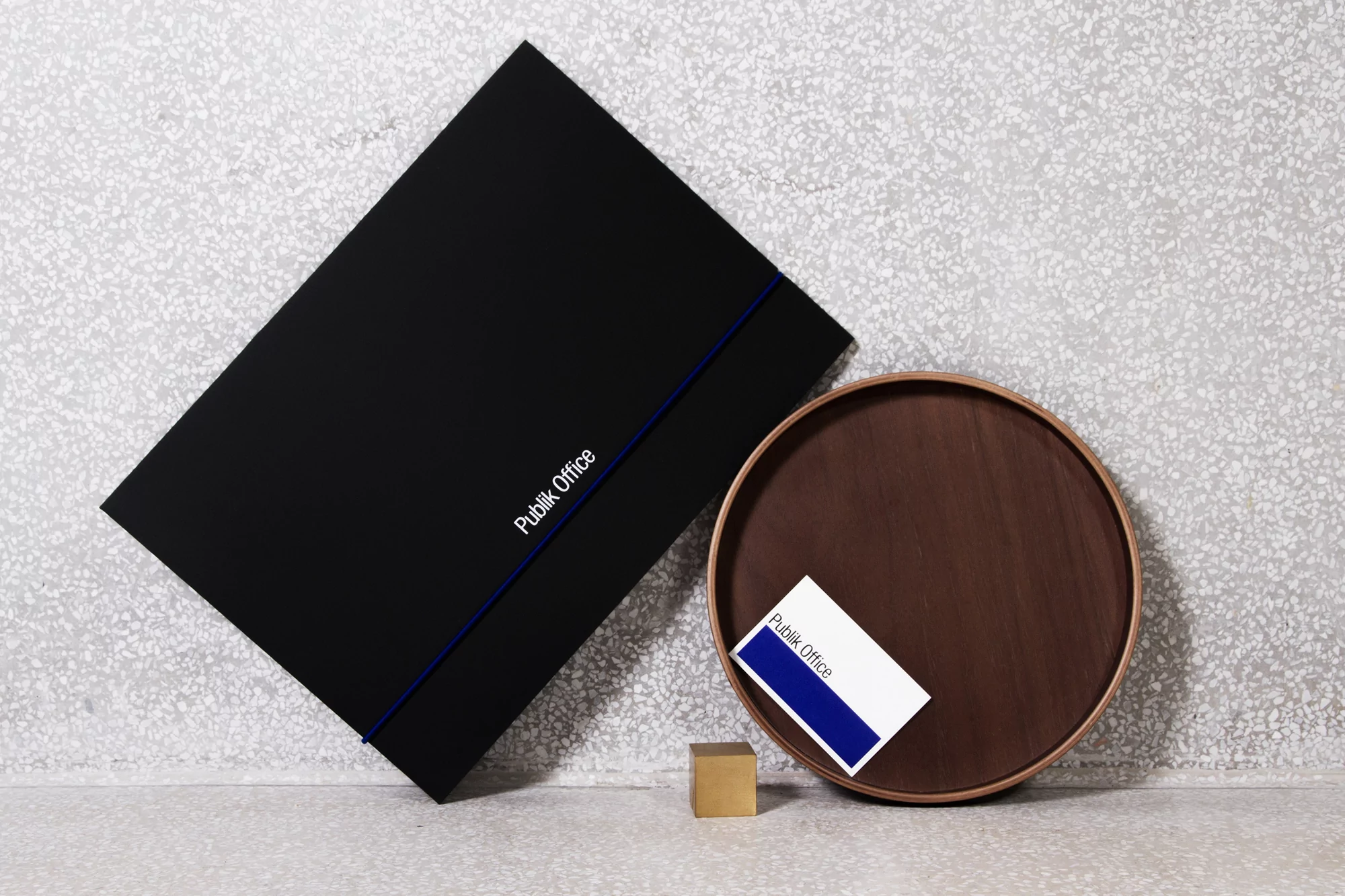 Elevated view of a black presentation folder with white text and a thin blue stripe next to a business card on a small wood tray.