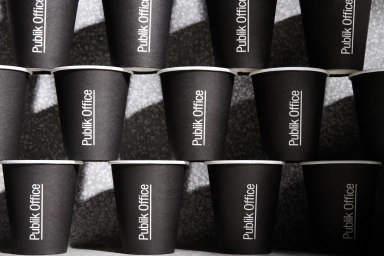 A neat stack of black paper coffee cups featuring the Publik Office brand logo on a speckled textured surface