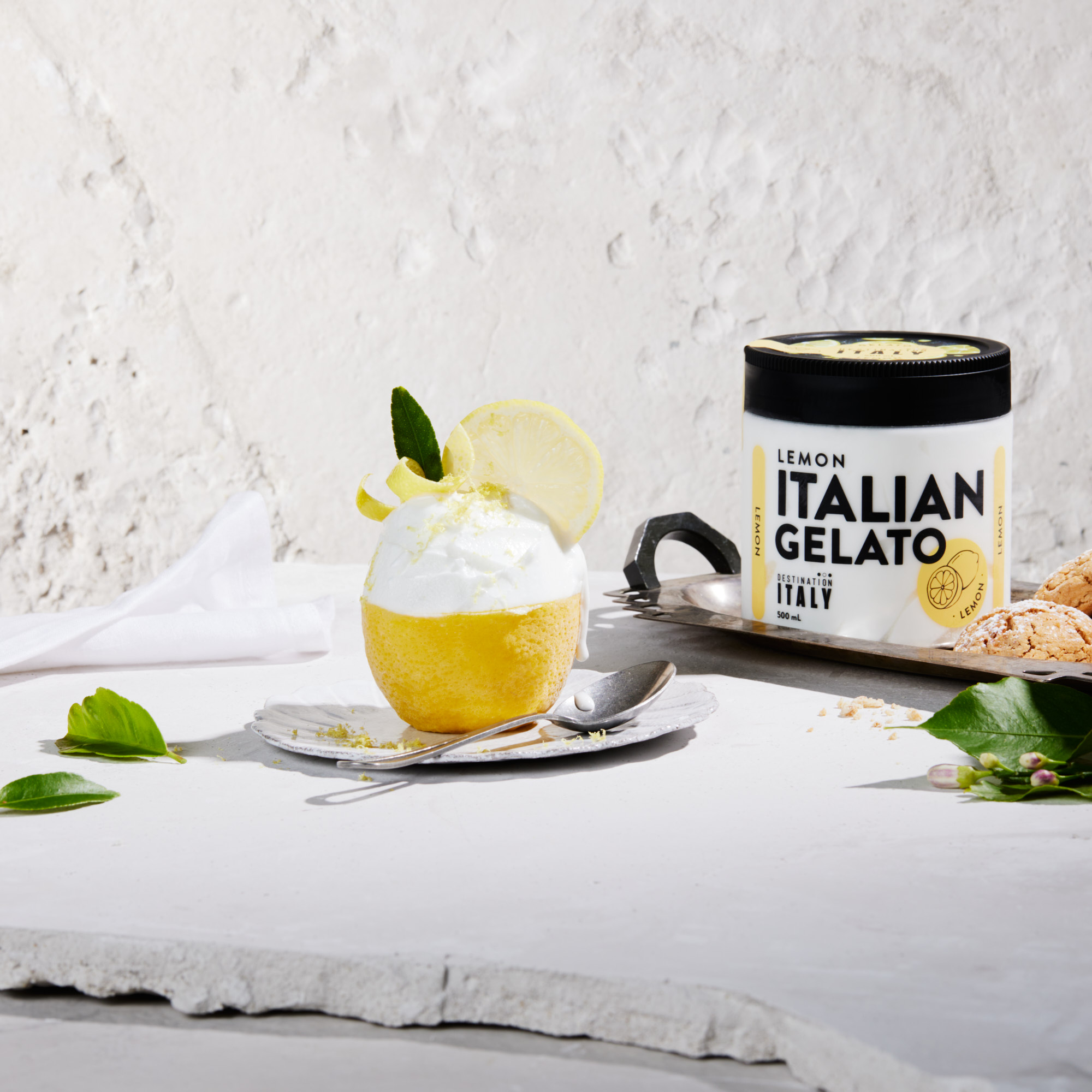 Lemon Italian gelato in a lemon rind cup on a silver tray with a jar of gelato, set against a textured off-white background.