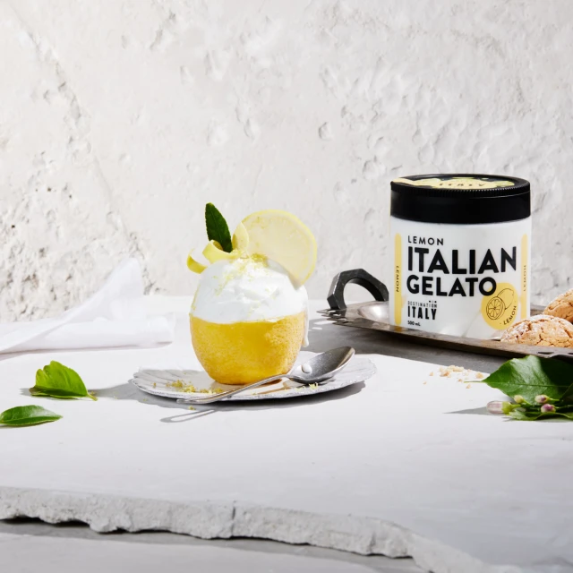 Lemon Italian gelato in a lemon rind cup on a silver tray with a jar of gelato, set against a textured off-white background.