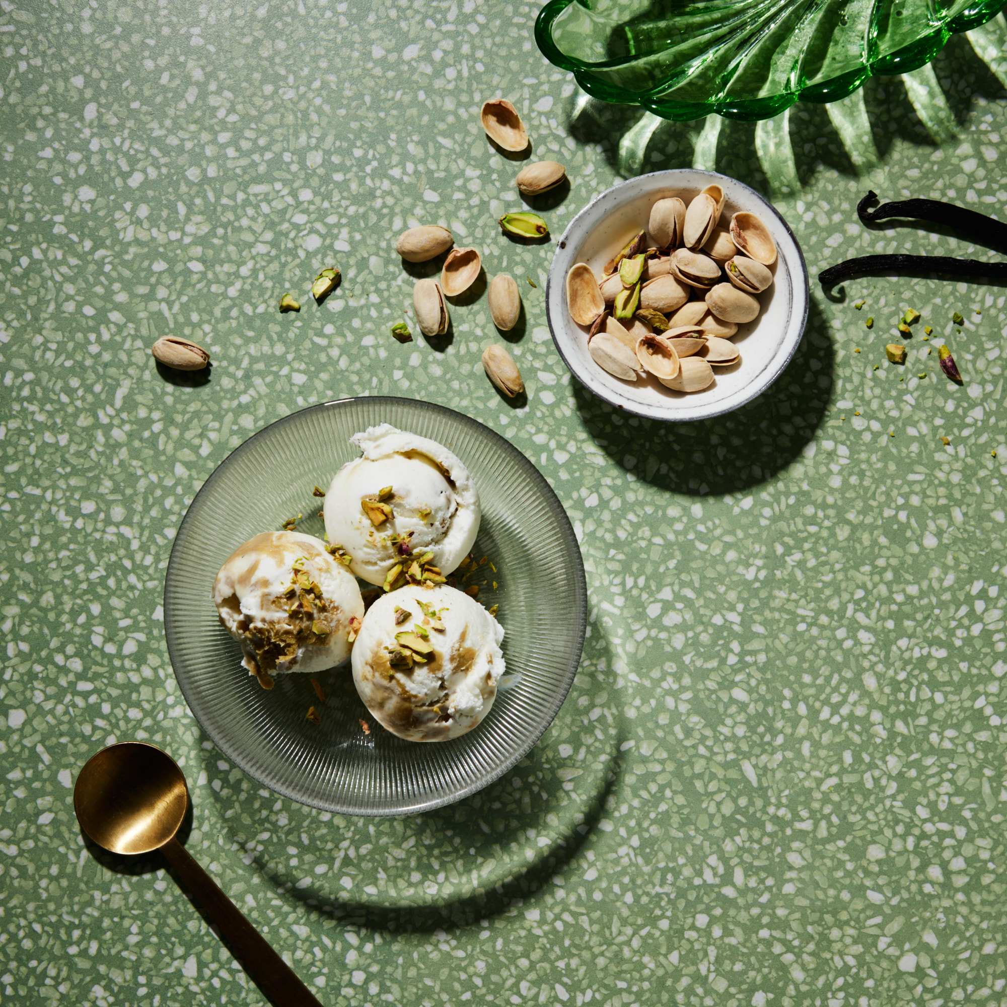 Overhead shot: three scoops of pistachio ice cream in a glass bowl on a green speckled surface, garnished with pistachios and spoon nearby.