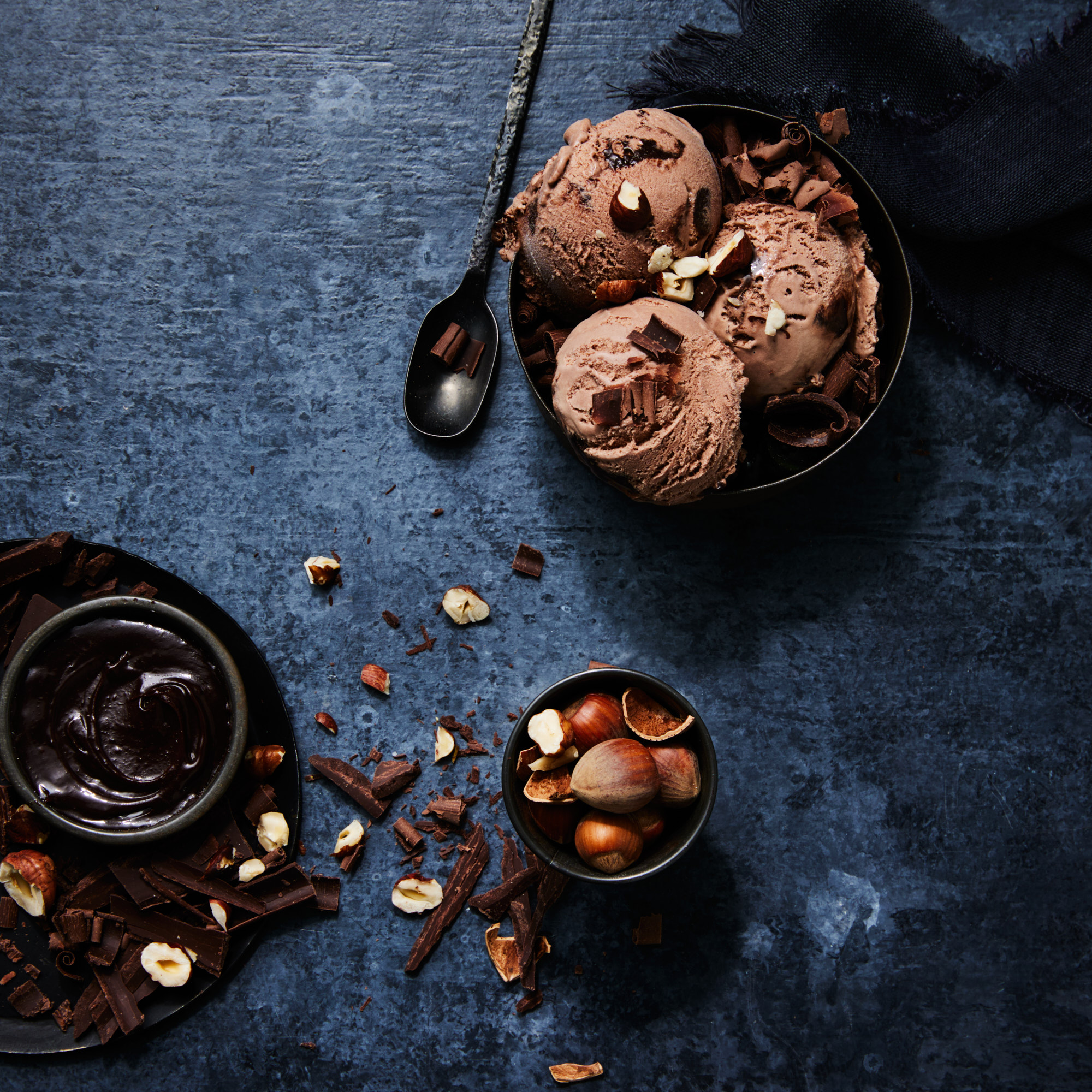 Overhead shot of chocolate ice cream scoops in a dark bowl, drizzled with nuts and chocolate shavings, atop a textured blue surface.