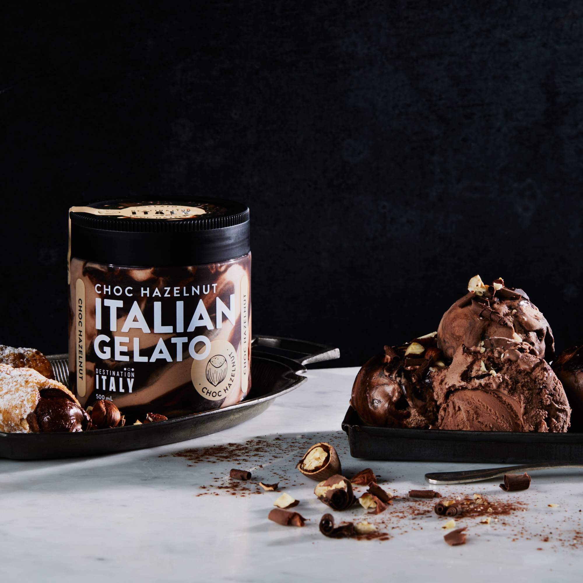 Chocolate hazelnut Italian gelato in a jar and plated scoops with chocolate cannoli on a marble surface against a dark background.