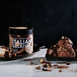 A tub of Choc Hazelnut Italian Gelato next to a tray of chocolate gelato scoops and pastry on marble