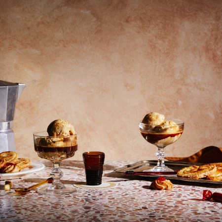 Aesthetic arrangement of affogato desserts in stemmed glasses, accompanied by pastries, coffee maker, and a small glass on a speckled table.