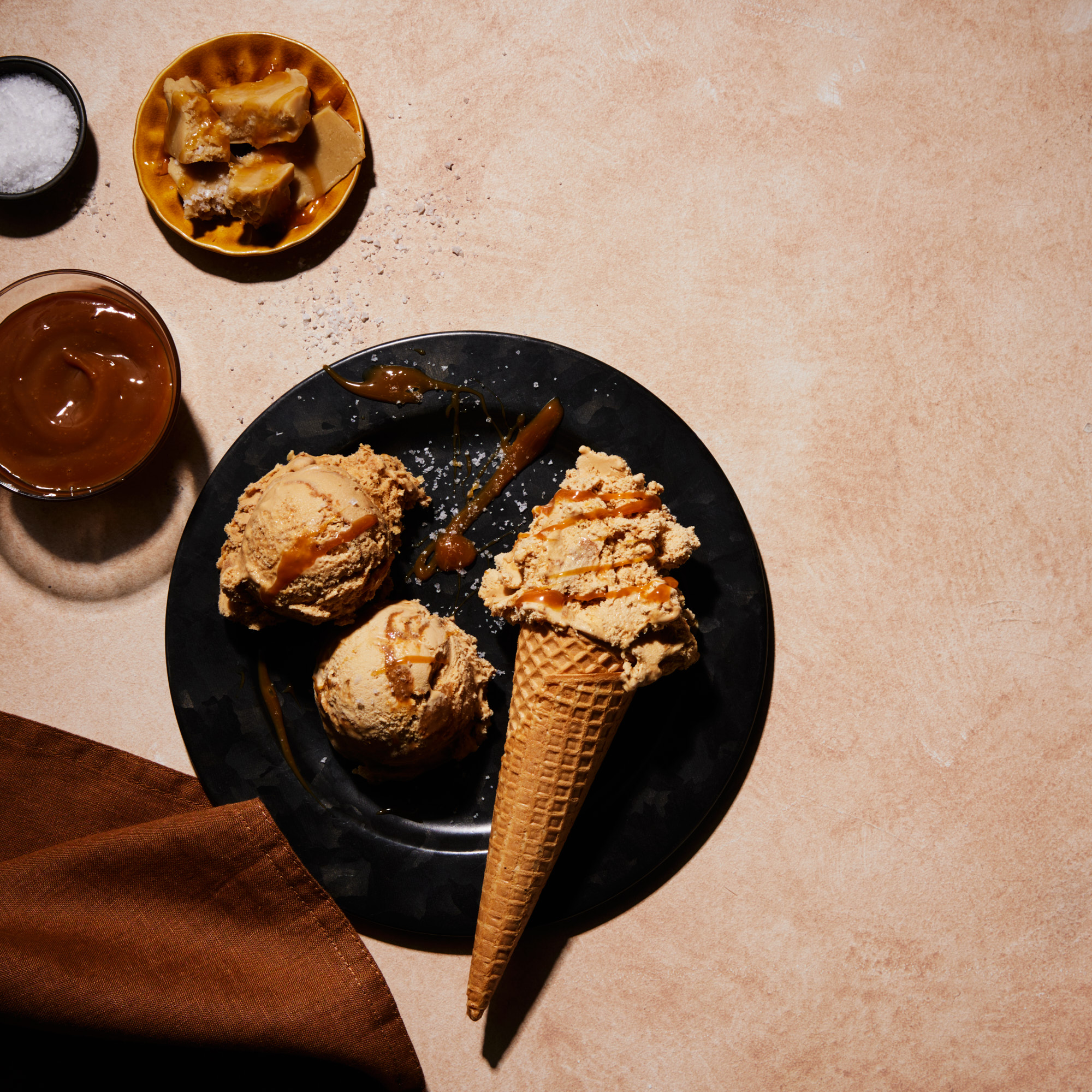 Eye-level shot of caramel ice cream on a black plate with caramel sauce, salt, and a brown napkin on a beige surface.