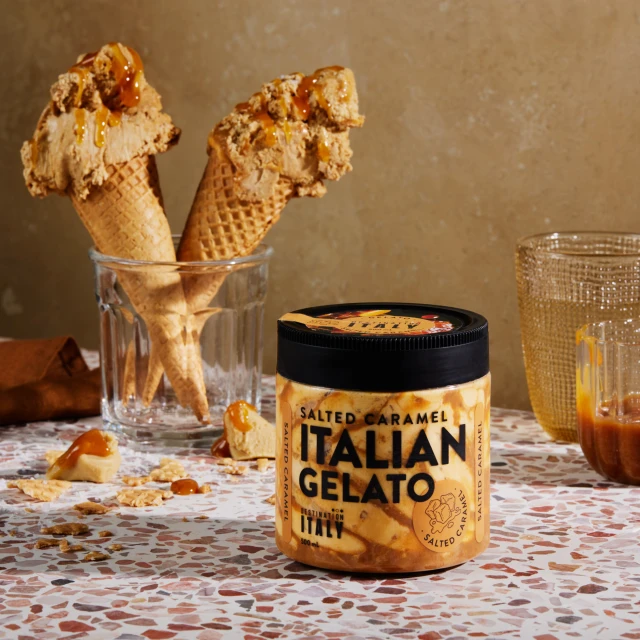 Two ice cream cones in a glass and a jar of Salted Caramel Italian Gelato sit on a terrazzo surface with a tan-colored background.