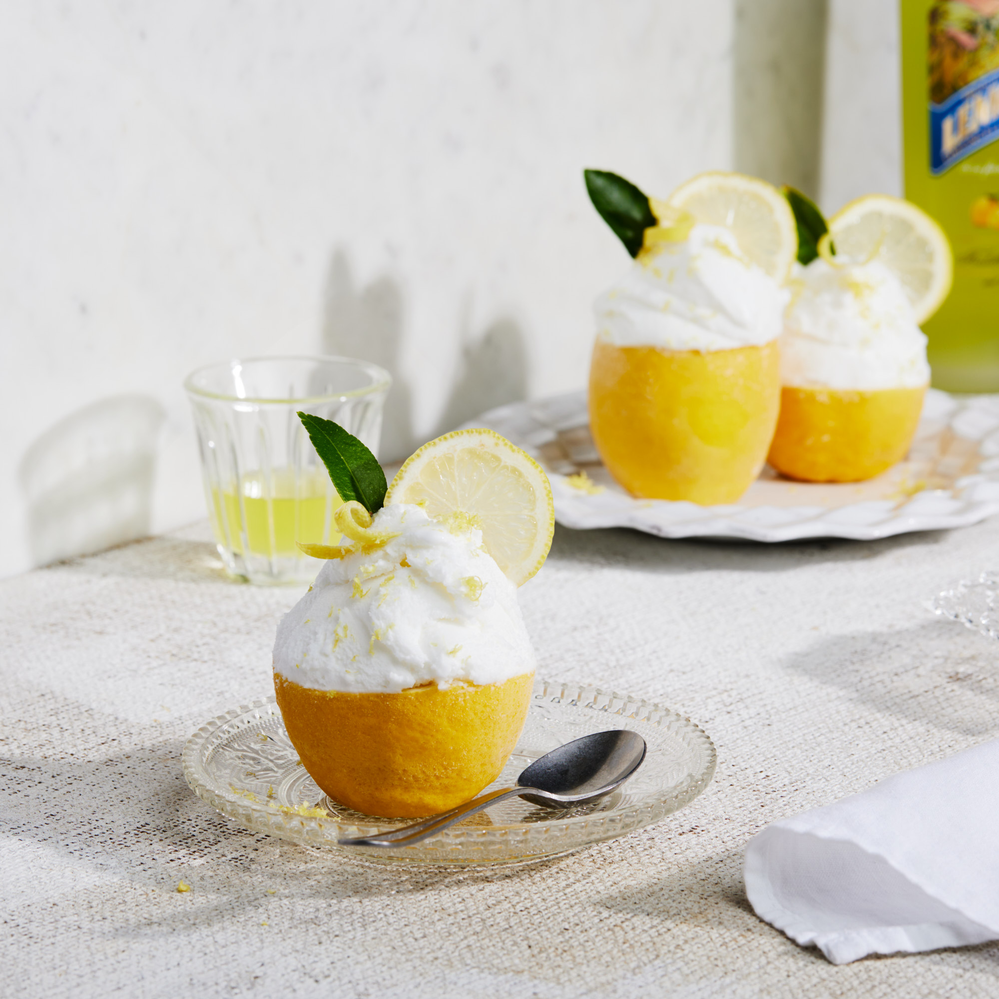 Lemon sorbet served in lemon peel bowls, topped with whipped cream and lemon slices, arranged on a table with a glass and bottle.