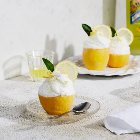 Lemon sorbet served in lemon peel bowls, topped with whipped cream and lemon slices, arranged on a table with a glass and bottle.