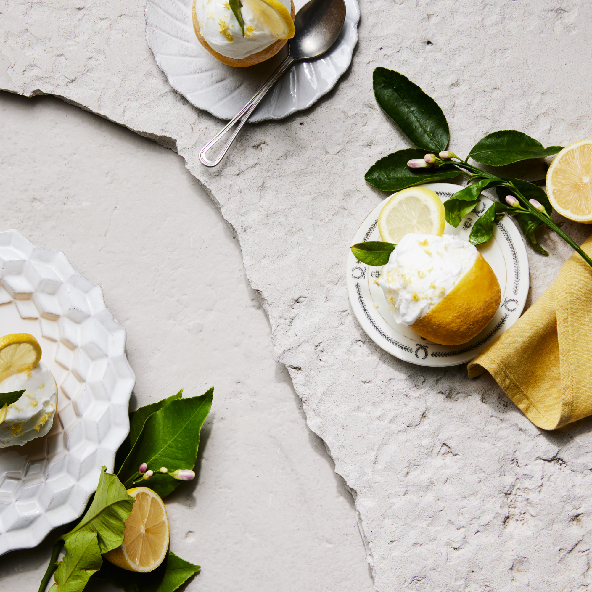 Overhead shot featuring lemon dessert cups on ornate plates, alongside lemon halves and a yellow napkin on a textured stone surface.