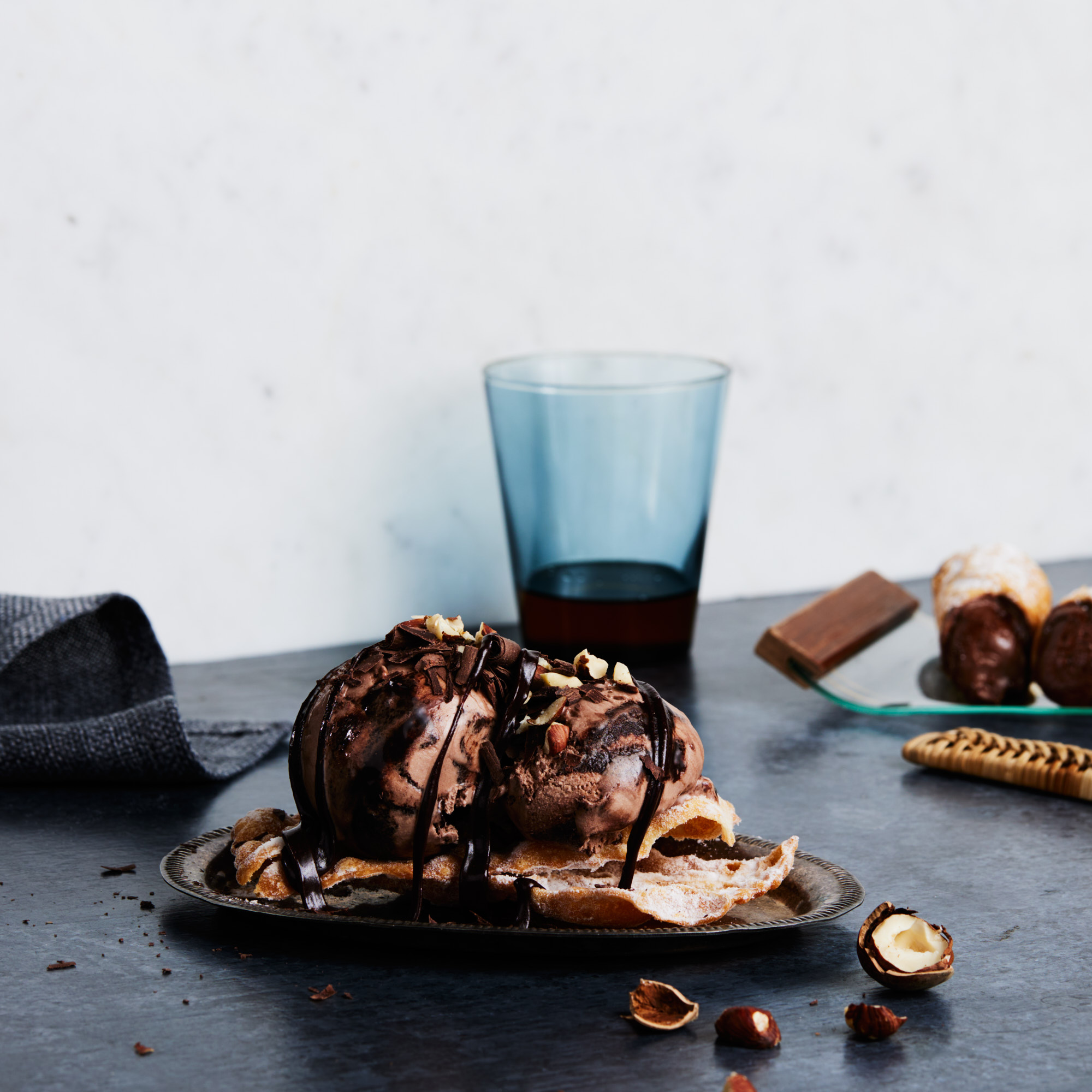 Two scoops of chocolate ice cream with chocolate syrup and chopped nuts, served on a plate with a drink and chocolates nearby.