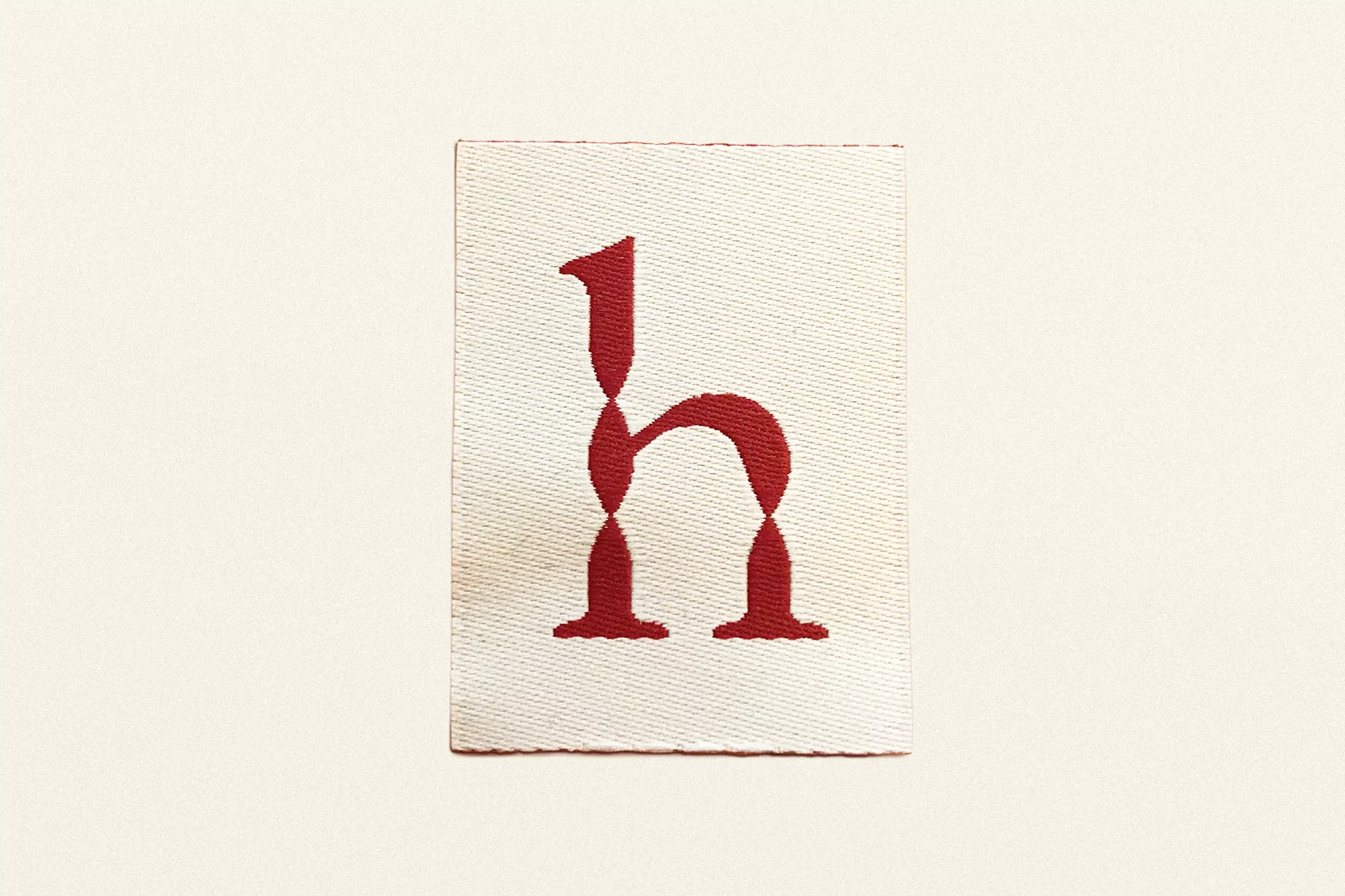 Fabric label with the letter h embroidered in red, set against an off-white backdrop. The tag has a woven texture.