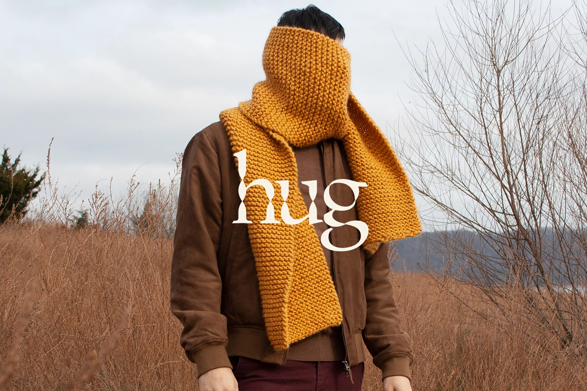 Person in a brown jacket and maroon pants wearing a yellow knitted scarf covering their face in a field of tall brown grasses.