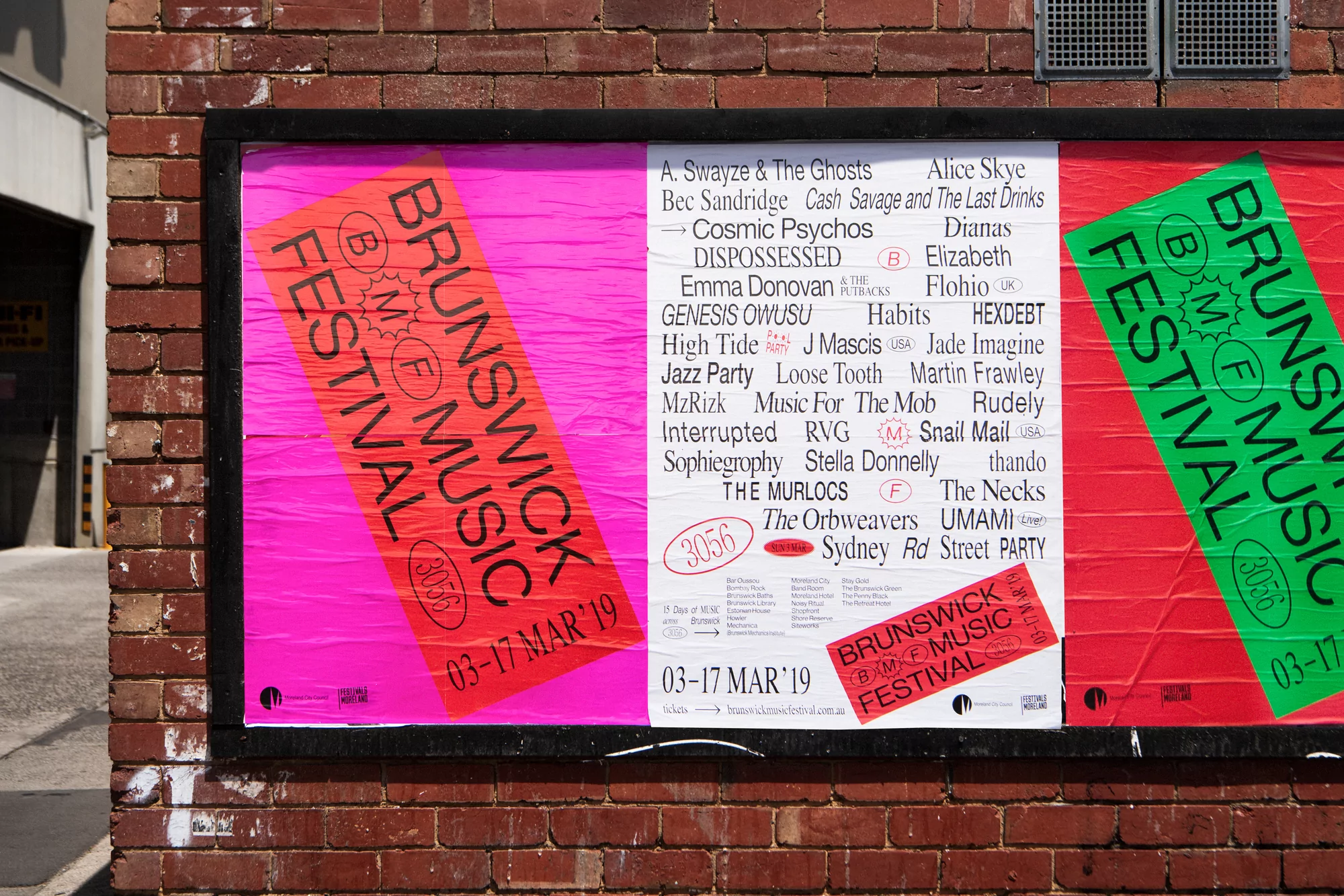 Outdoor posters attached to a red brick wall, promoting the Brunswick Music Festival. Pink and green colors with printed text on the posters.