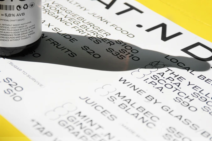 Close-up photo of a beer bottle and a menu on a bright yellow surface, highlighting the drink selection and pricing.