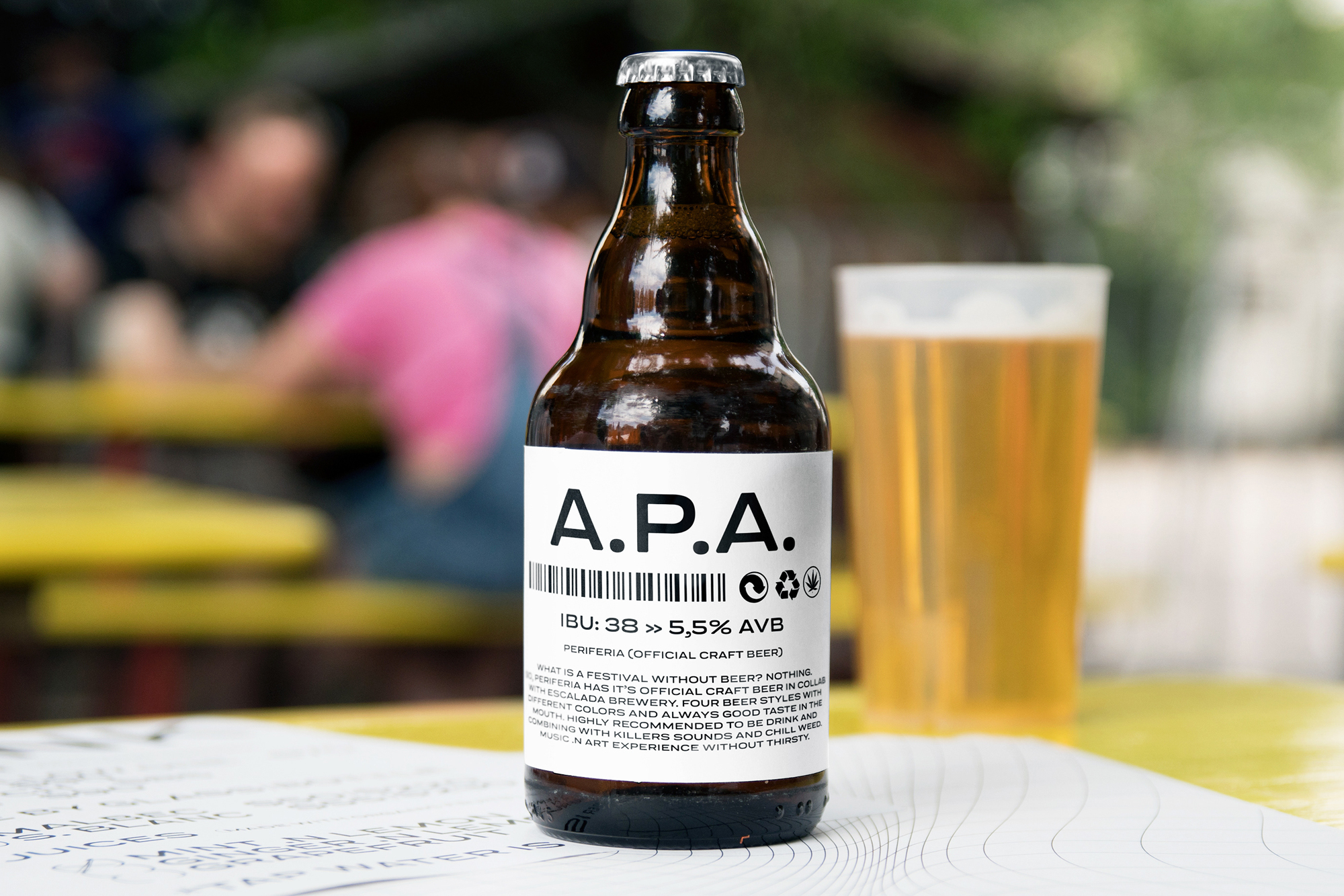 Brown glass beer bottle with a white label and silver cap sits on a yellow table with a pint of beer in the background.