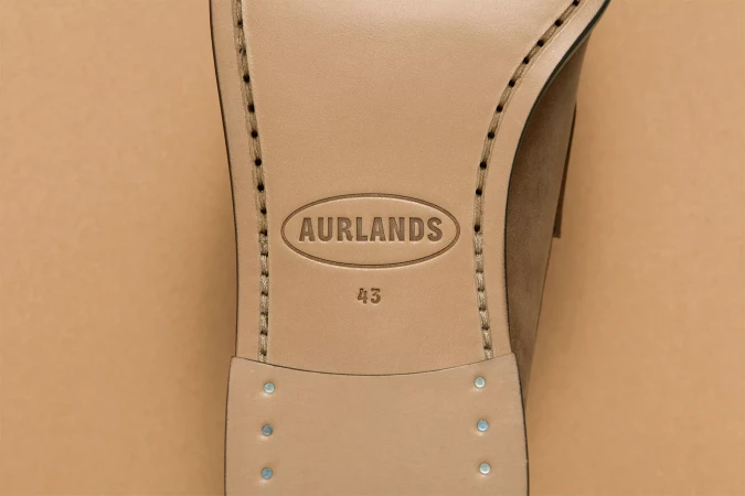 Close-up studio shot of a tan-colored leather dress shoe sole featuring stitching details and the 'Aurlands' logo against a tan backdrop.
