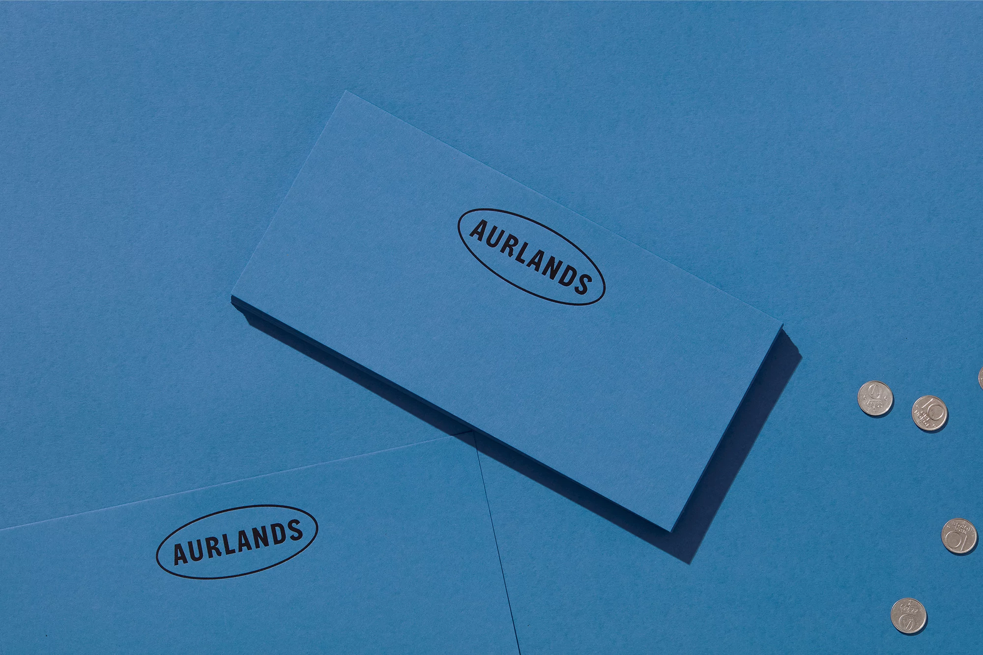 Overhead shot of blue stationery cards with Aurlands logo and scattered coins on a matching blue background creating a minimal composition.