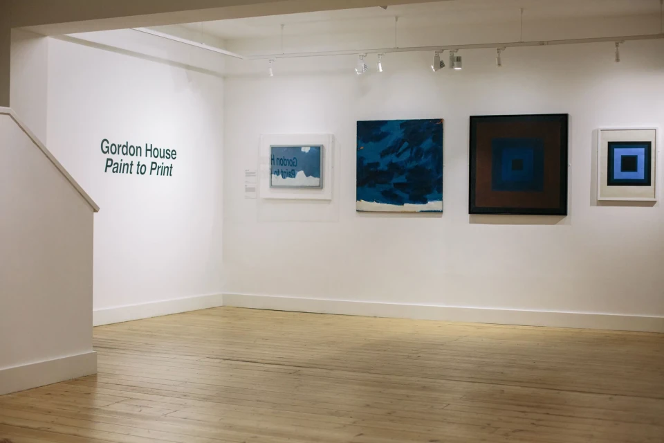 Art gallery featuring abstract paintings with square motifs in shades of blue, brown, and white hanging on white walls, indoor setting.