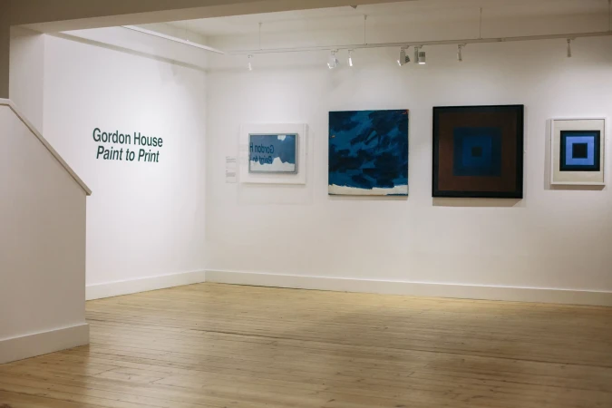 Art gallery featuring abstract paintings with square motifs in shades of blue, brown, and white hanging on white walls, indoor setting.