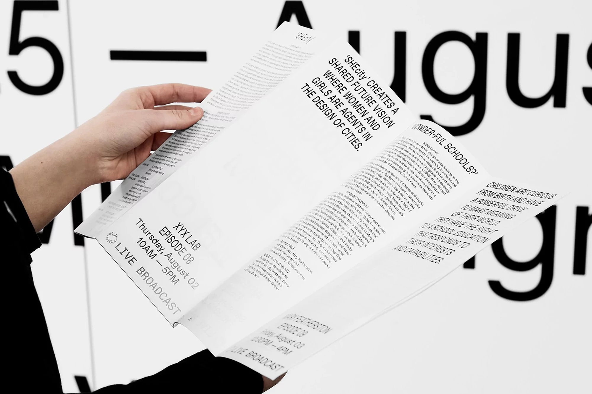 Close-up of a person holding an open booklet with text, standing in front of a wall with large black text; indoor setting.