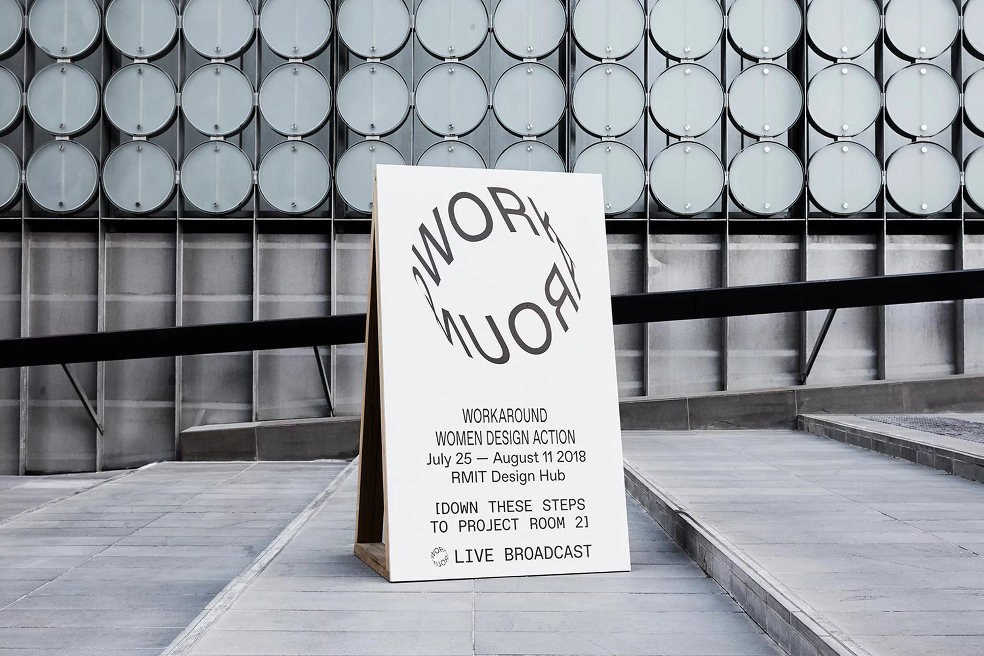 Promotional sign for the 'Workaround Women Design Action' event, featuring graphic typography and minimalist design, standing in an outdoor urban setting.