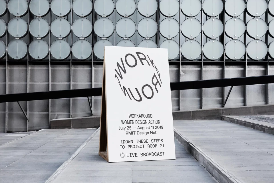 Promotional sign for the 'Workaround Women Design Action' event, featuring graphic typography and minimalist design, standing in an outdoor urban setting.