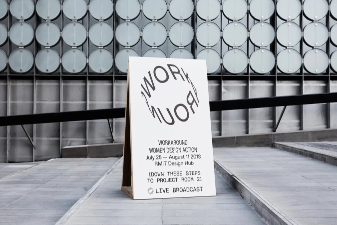 Promotional sign for the 'Workaround Women Design Action' event, featuring graphic typography and minimalist design, standing in an outdoor urban setting.