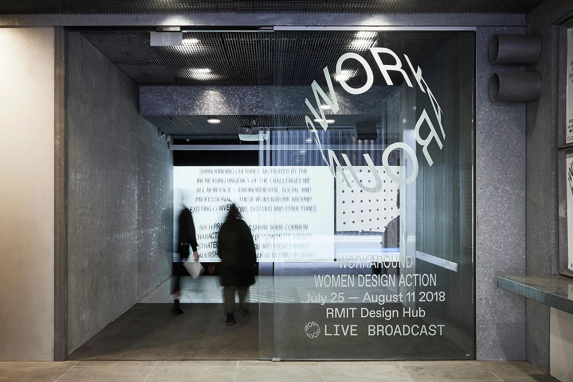 Architectural shot of a glass door with text at RMIT Design Hub, featuring two blurred figures walking into an exhibition space.