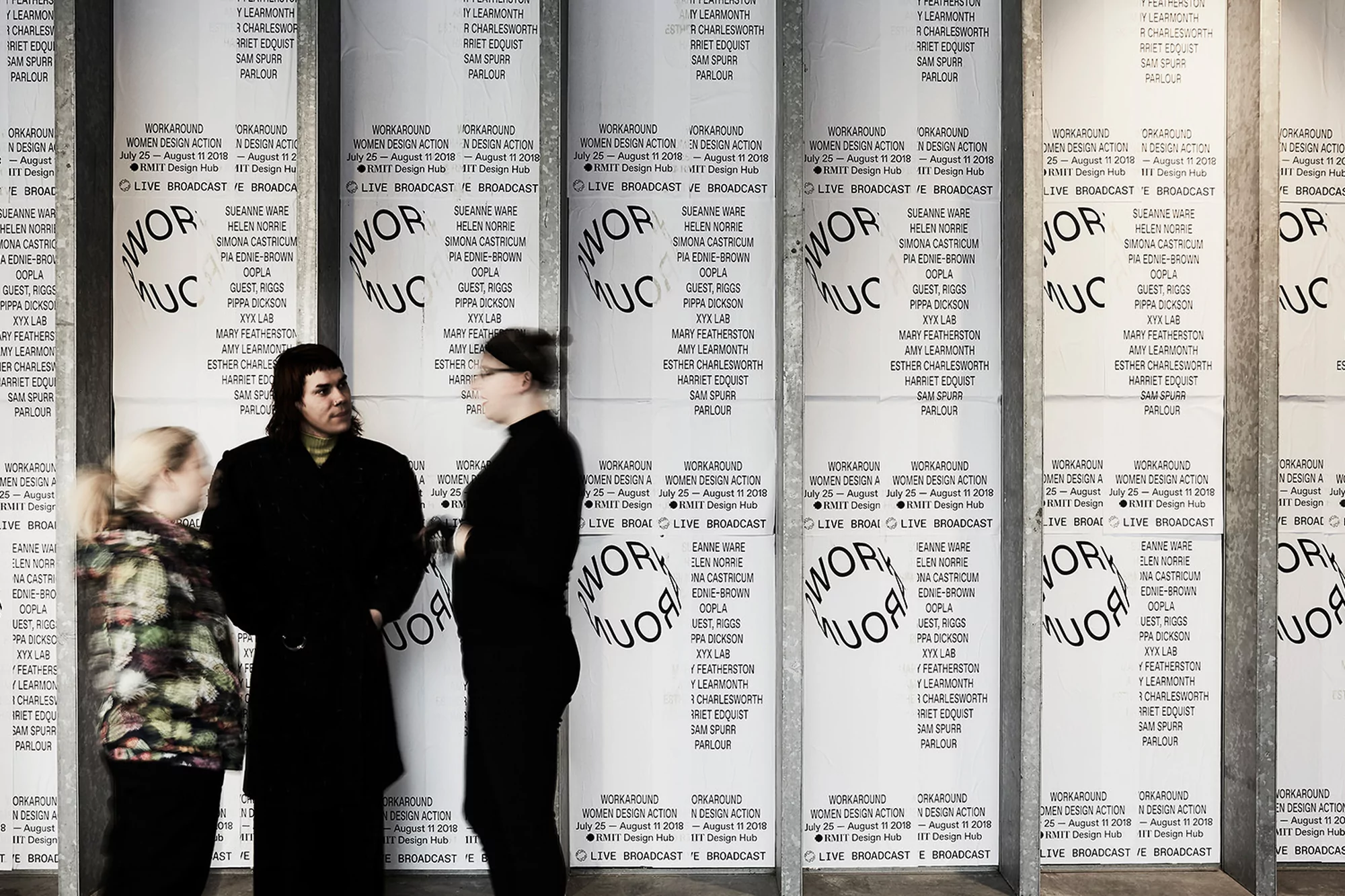 Three people stand in front of large posters with text and circular design. The person in the middle is facing the camera.