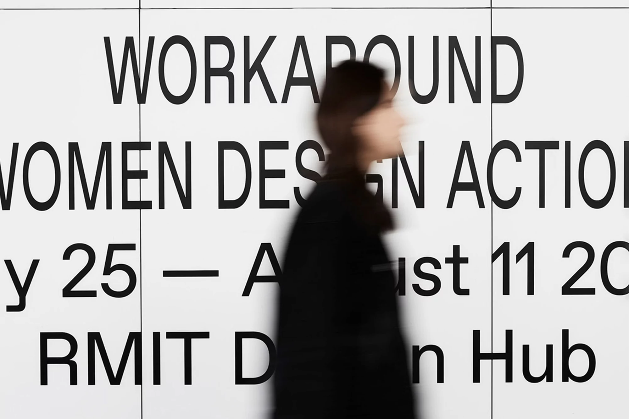 Blurred person walks past large format poster with text 'WORKAROUND WOMEN DESIGN ACTION July 25 – August 11 2024 RMIT Design Hub'.