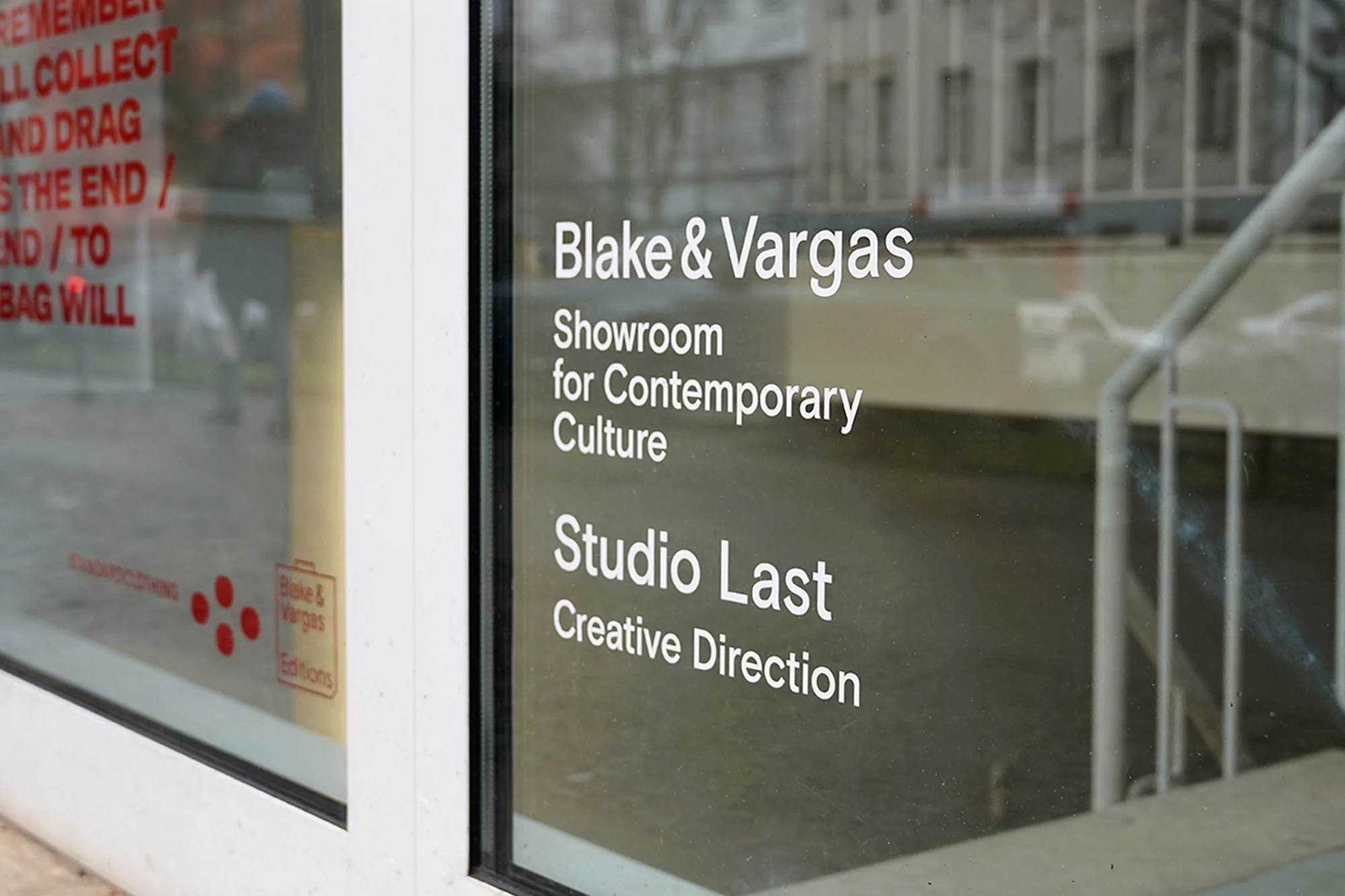 Exterior shot of a showroom window with white text reading 'Blake & Vargas' and 'Studio Last,' reflecting an urban scene.
