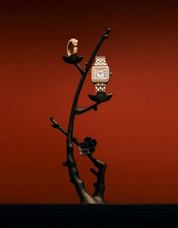 Gold watch and ring are displayed on a black sculptural tree against an orange backdrop, highlighting jewelry and timepieces.