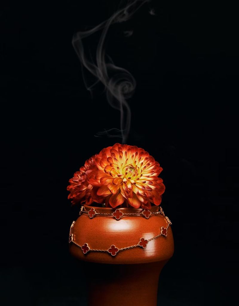 High-angle studio shot of an orange flower arrangement and cloverleaf-pattern gold necklace on a terra cotta vase against a black background.