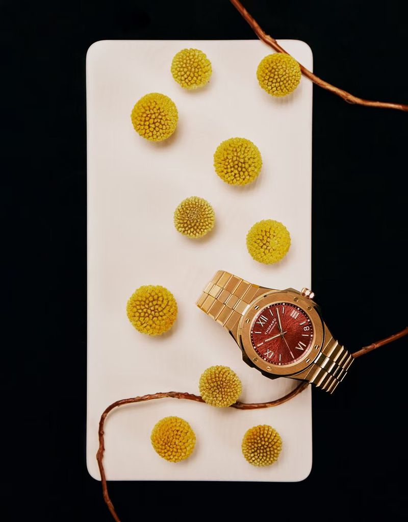 Elegant gold wristwatch with a burgundy face on a white surface surrounded by yellow flowers and copper branches against a dark background.