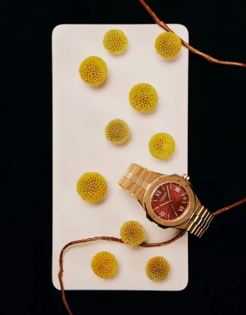 Elegant gold wristwatch with a burgundy face on a white surface surrounded by yellow flowers and copper branches against a dark background.