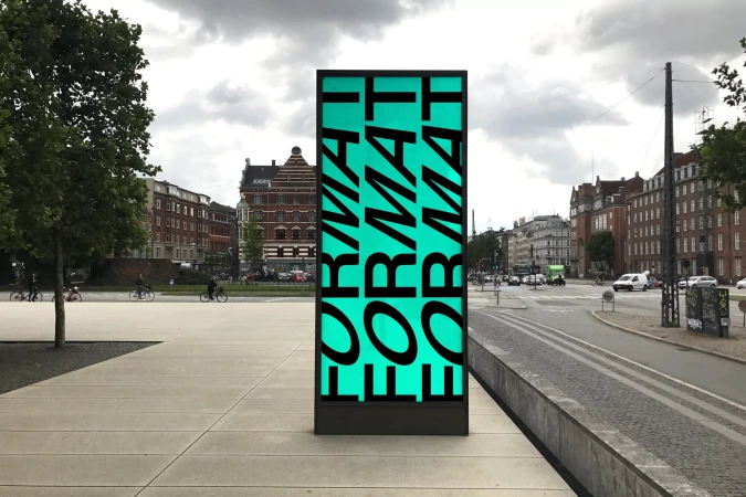 Vertical sign with the word 'FORMAT' in black letters on a green background, standing on a concrete surface in an urban environment.