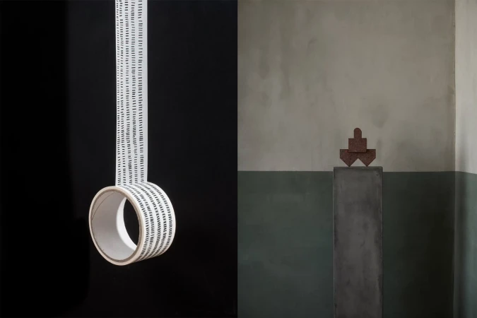 Abstract studio shot of white tape roll with black dashes suspended in mid-air, with minimalist sculpture on a plinth against green wall.